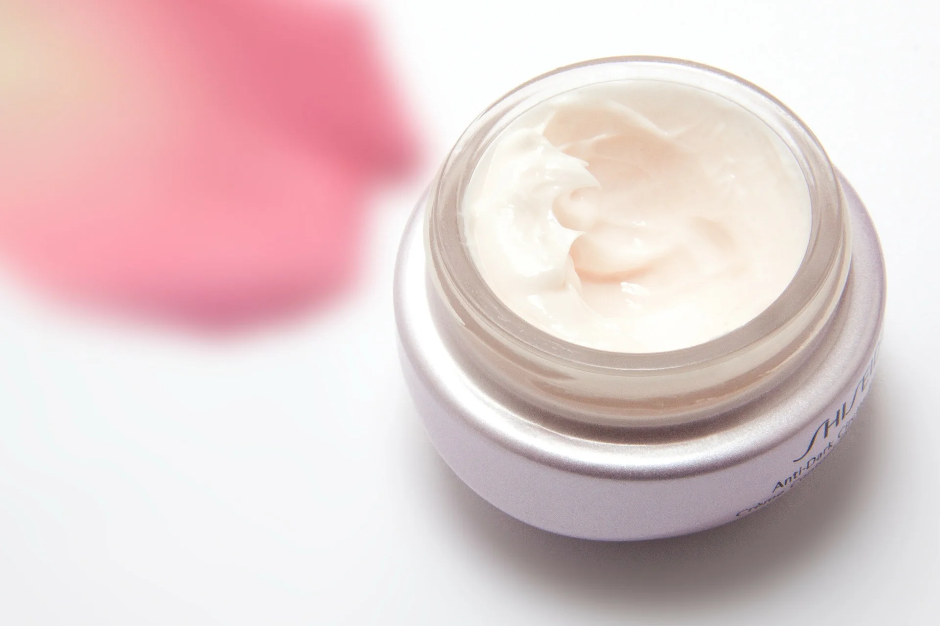 Open jar of skincare cream on a white surface with a blurred pink flower beside it.